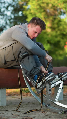 un tipo sentado en un tronco en un parque, con una camisa gris de mangas largas y pantalones de carga, centrado en ajustar las correas de muelle de primavera unidas a los pies, rodeado de árboles verdes, con un ángulo de cámara bajo
