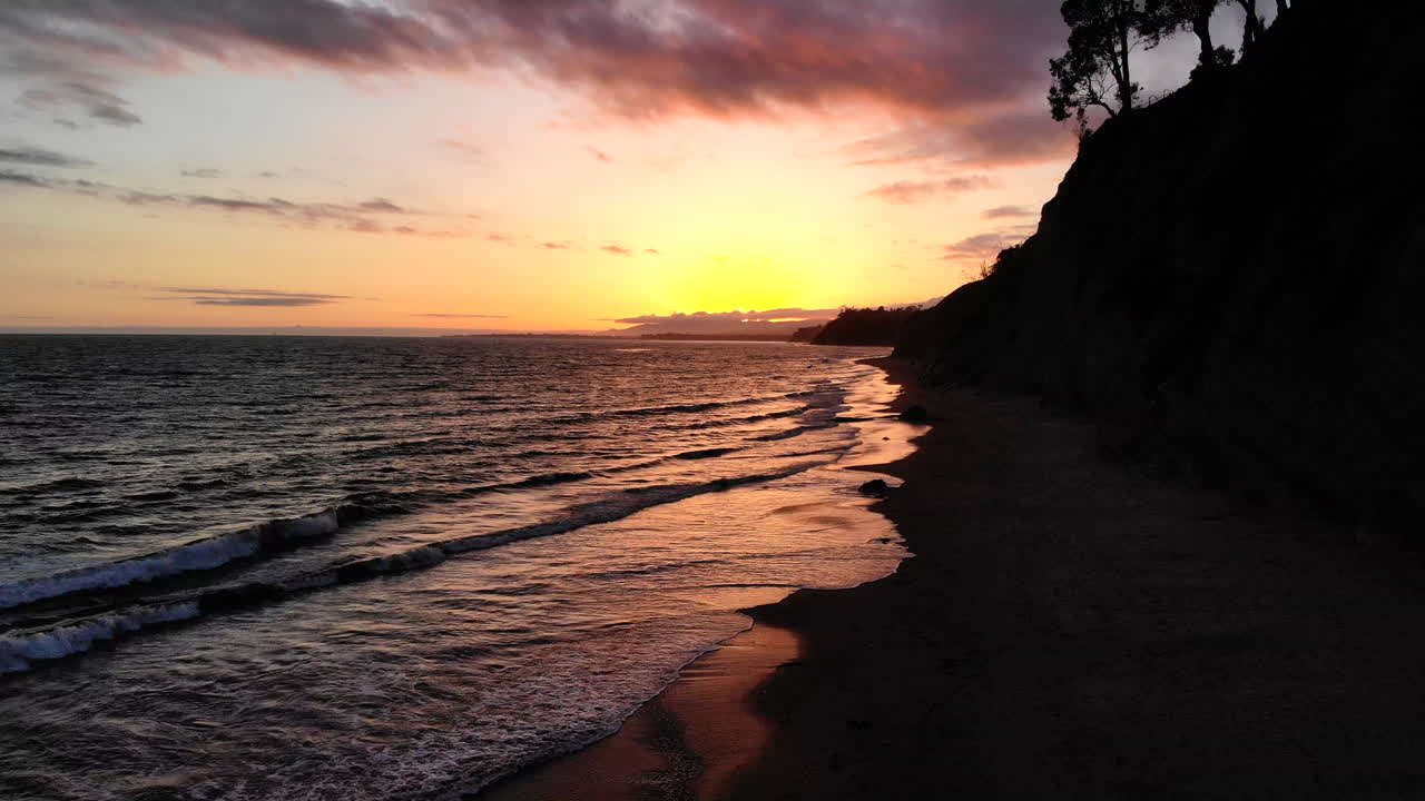 toma aérea de un cielo de puesta de sol sobre el océano y reflejos en la playa de arena húmeda cerca de los acantilados de la costa de santa barbara, california