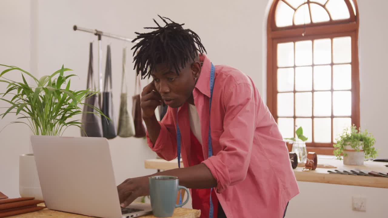 Heureux artisan afro-américain avec des dreadlocks passant un appel téléphonique et utilisant un ordinateur portable dans l'atelier.