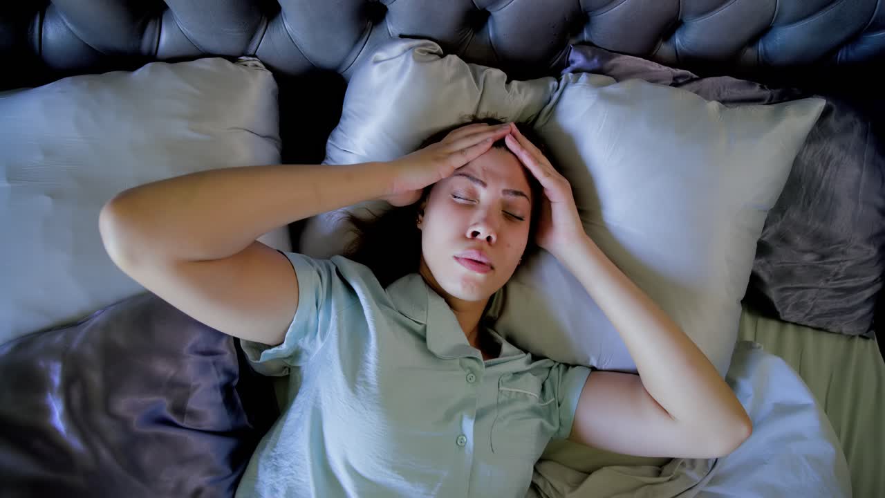 Donna afroamericana a letto che si sveglia al sole, si massaggia le tempie cercando sollievo dal dolore con i cuscini, indossando un top color menta.