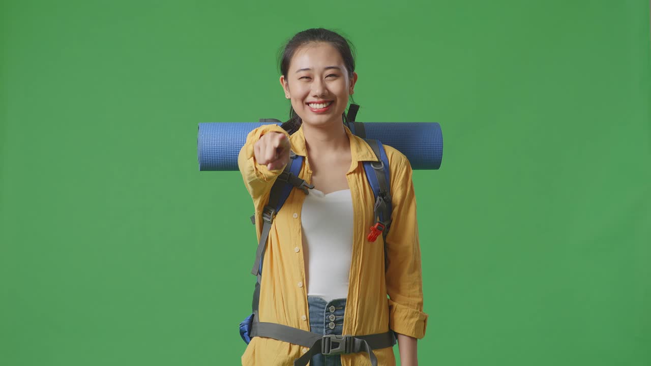 excursionista asiática con mochila de montañismo sonriendo, tocando su pecho y señalando a la cámara mientras está de pie en el fondo de la pantalla verde en el estudio