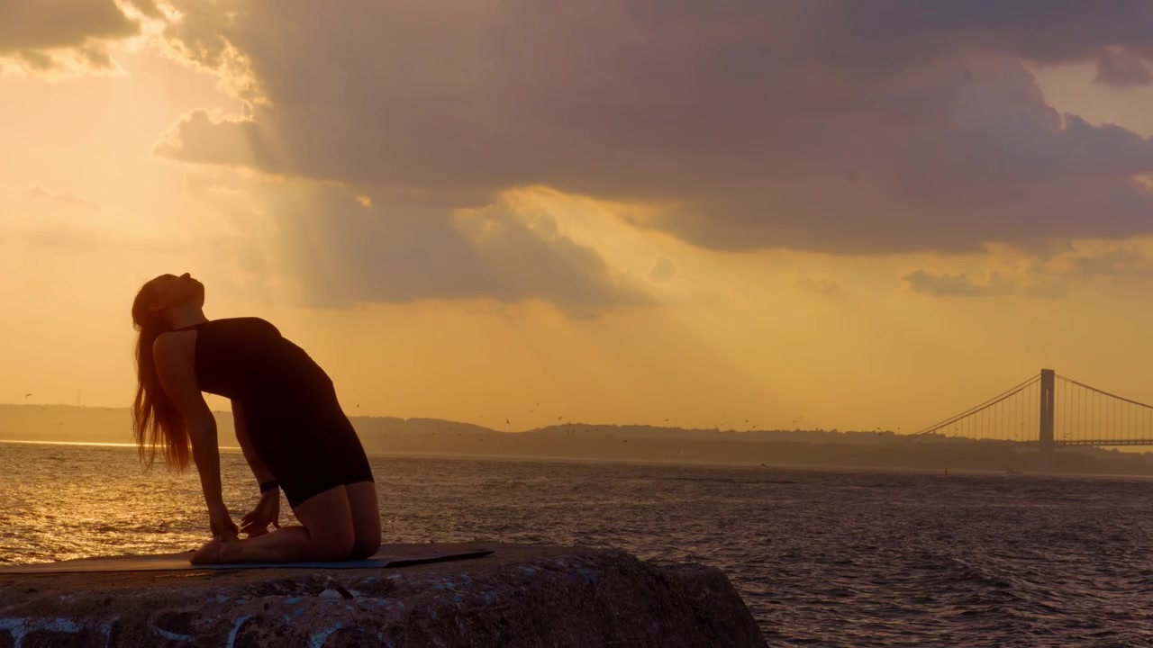 Mulher com mãos em posição de oração em pose de yoga ajoelhada ao anoitecer. Silhueta de perfil com palmas das mãos levantadas em cais de pedra, Ponte Verrazzano-Narrows, céu de pôr do sol amarelo.