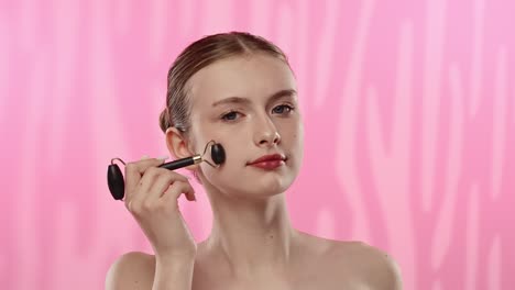 young woman using facial roller on clear skin in spa environment a young woman applies a facial roller to her face in a serene spa setting with a soft pink backdrop promoting skincare routine