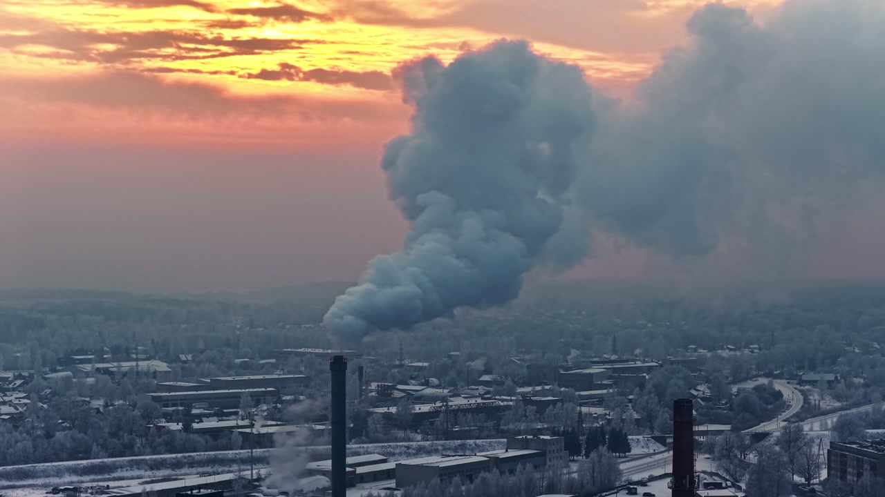 Aerial parallax reveals factory chimneys billowing thick white fumes against pink orange sunset over snow-covered Jelgava city buildings and roads in winter, Latvia