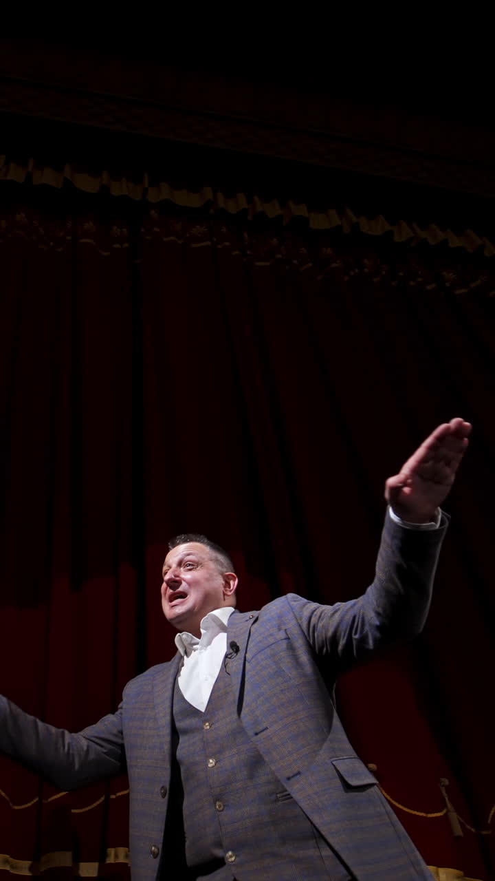 Theatrical man on stage. Performer in a suit moving on stage on red curtains background. Professional actor performing in theater. View from below. Vertical video