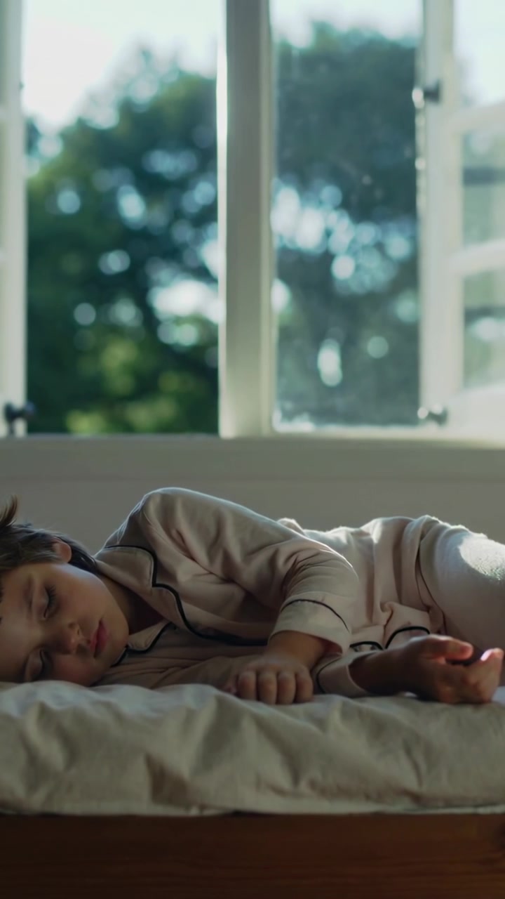 縦型動画：開いた窓の下のベッドで、服を着たまま昼寝をする子供、日差しが移り変わる
