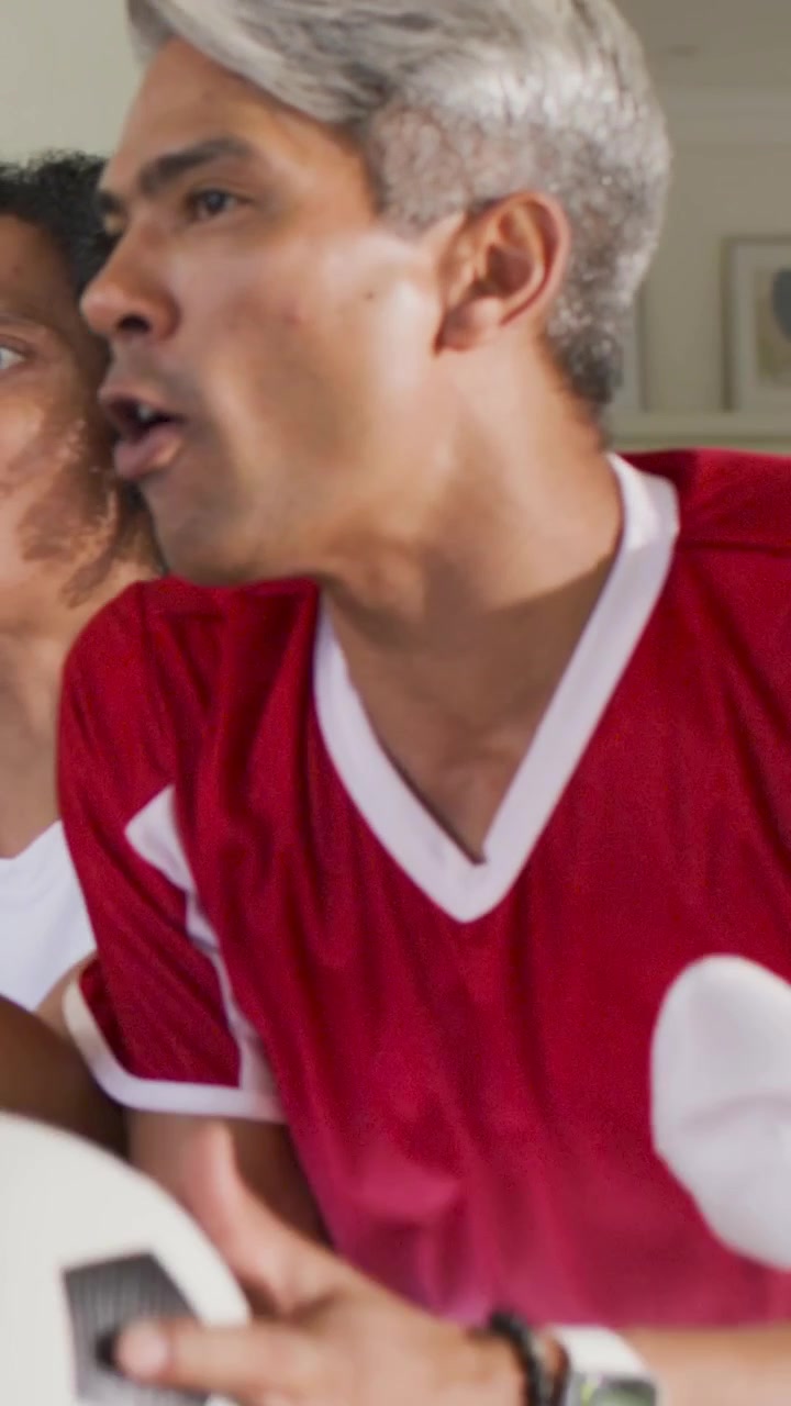 Video verticale: Uomo con maglia rossa che urla sul divano mentre un pallone da calcio gli passa vicino al viso.