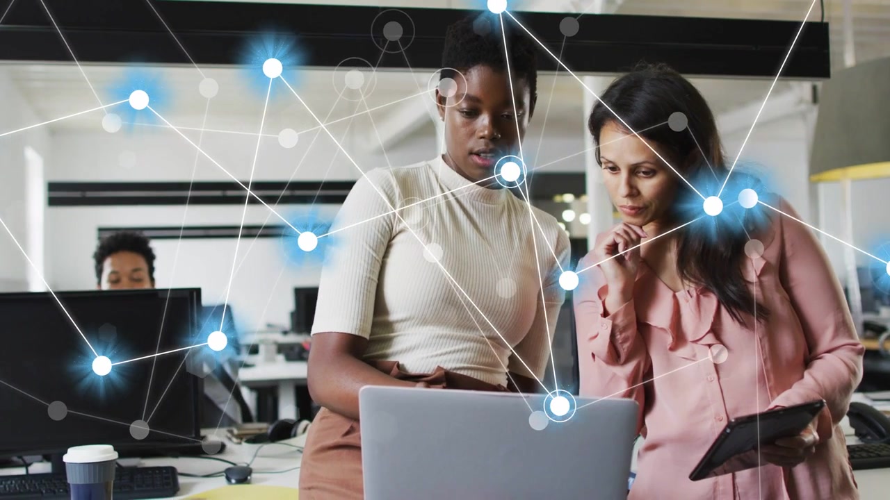Two women exchanging cues, leaning over laptop and tablet, network overlay growing, analyzing data