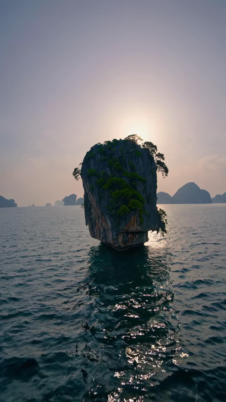Aerial video captures a solitary rock island at sunset, highlighting its lush greenery and dramatic