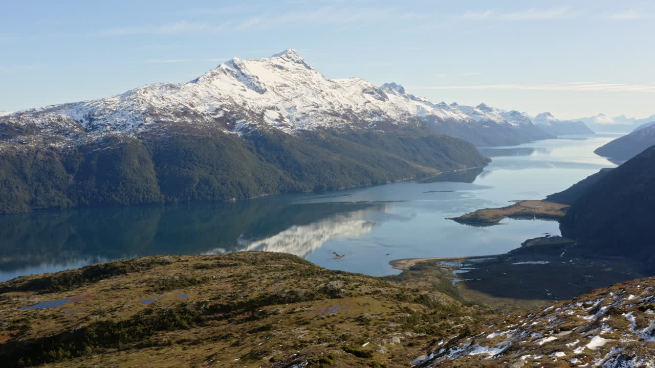 Snowcapped mountain range behind fjord inlet with green slopes and cold coastal winds, aerial panoramic establishing with reflection in water