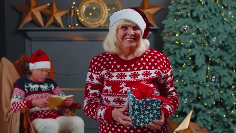 elderly grandmother in festive sweater presenting christmas gift box, smiling, looking at camera