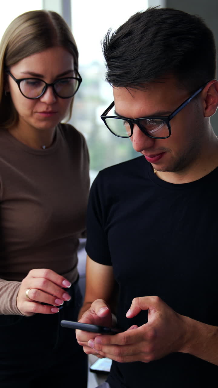Colleagues at office during the lunch break. Man holding a phone and woman comes up to help him. Panoramic windows at backdrop in blur. Vertical video