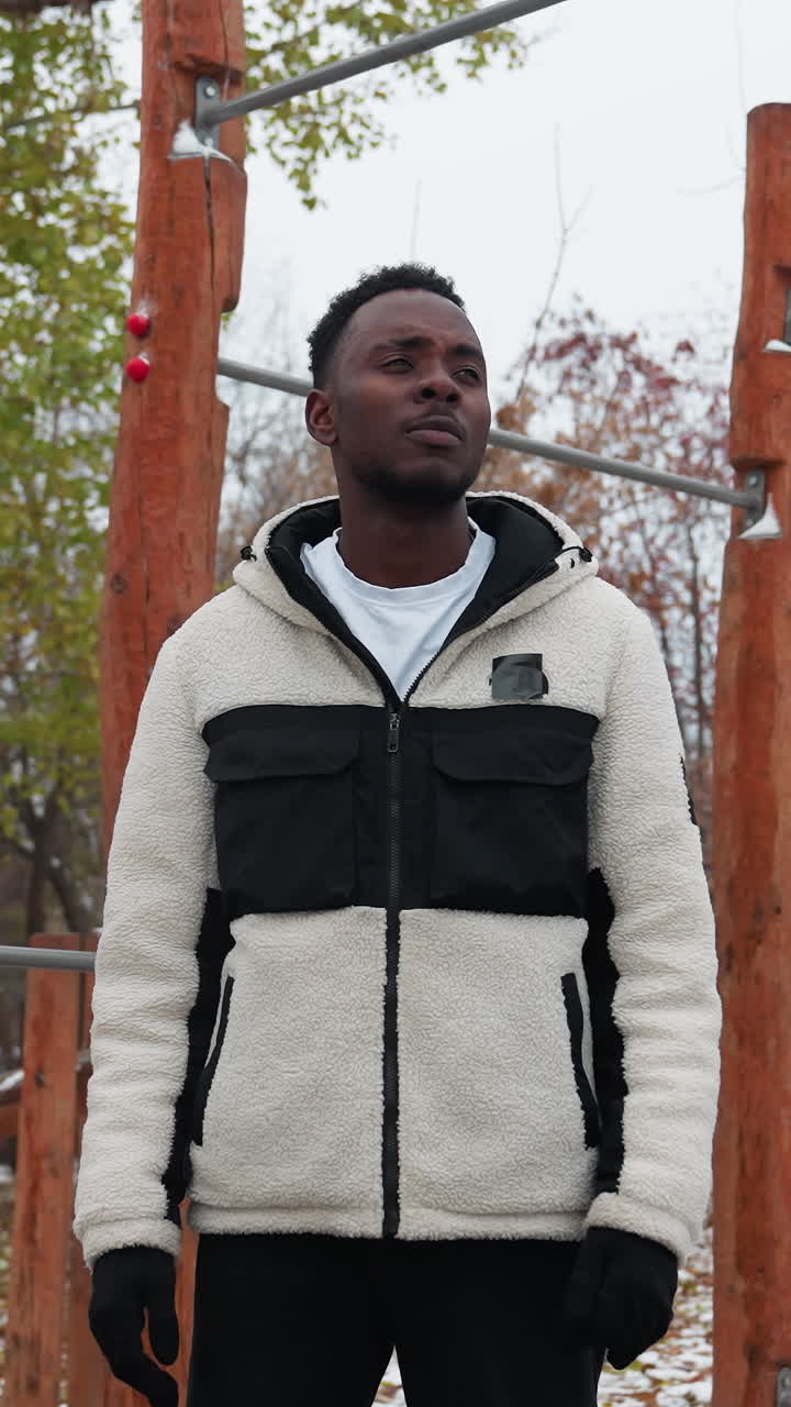 Boy standing still in outdoor workout park, gazing into distance with thoughtful expression, dressed in warm jacket, surrounded by trees, autumn leaves, snow patches, and fitness equipment