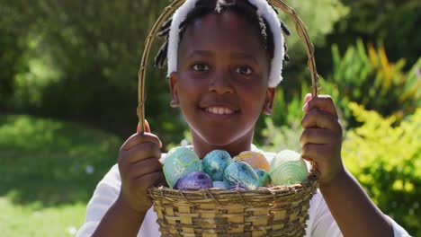 smiling african american girl wearing easter bunny ears holding basket of easter eggs in garden