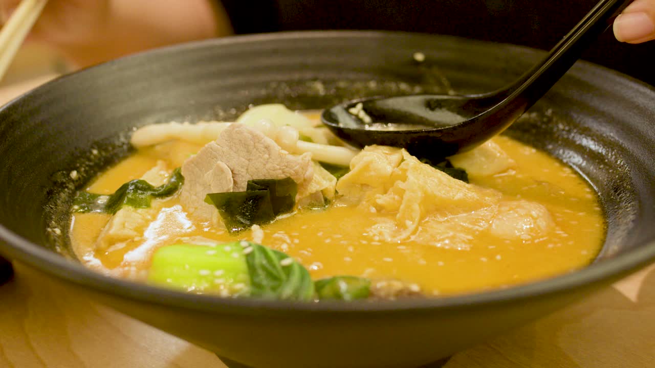 Person uses chopsticks and spoon to eat spicy noodle soup under warm indoor lighting, close-up