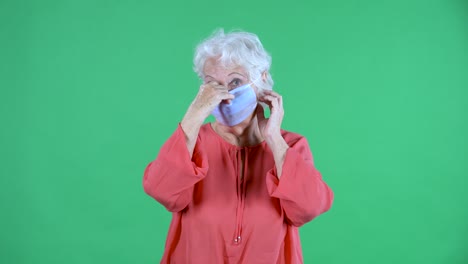 retrato mujer mayor mirando a la cámara y rputs en máscara médica. abuela de cabello gris en blusa roja en la pantalla verde en el estudio. cerca