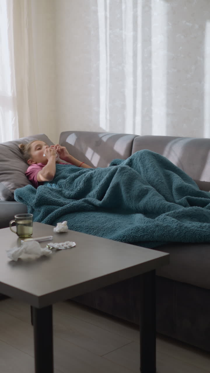 Bambino malato sdraiato sul divano, coperto da una coperta, che si sistema, circondato da fazzoletti sparsi, sintomi influenzali visibili con fazzoletti, medicine e bevanda sul tavolo in soggiorno