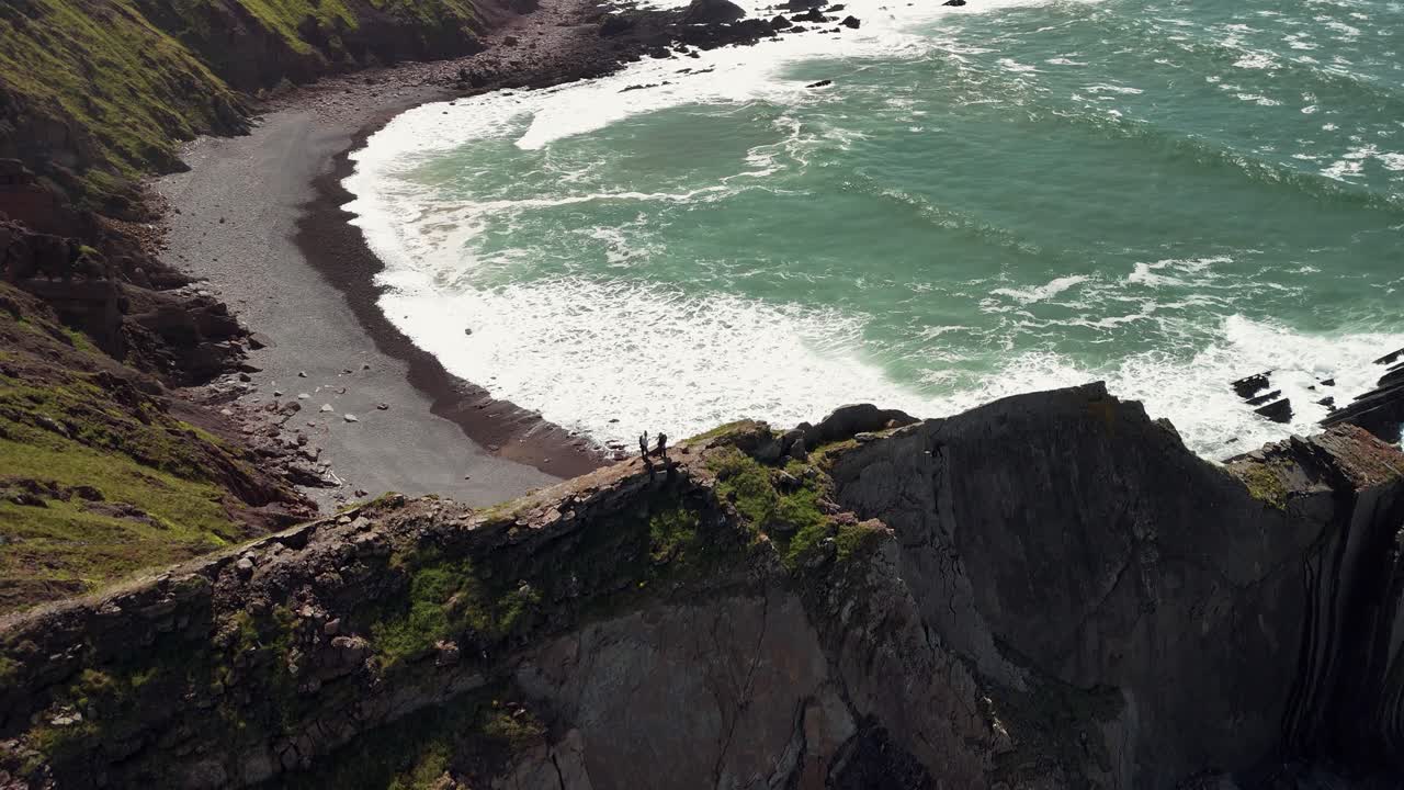 Slow, pull up aerial shot revealing a couple hiking on a cliff edge overlooking a wild, hidden beach