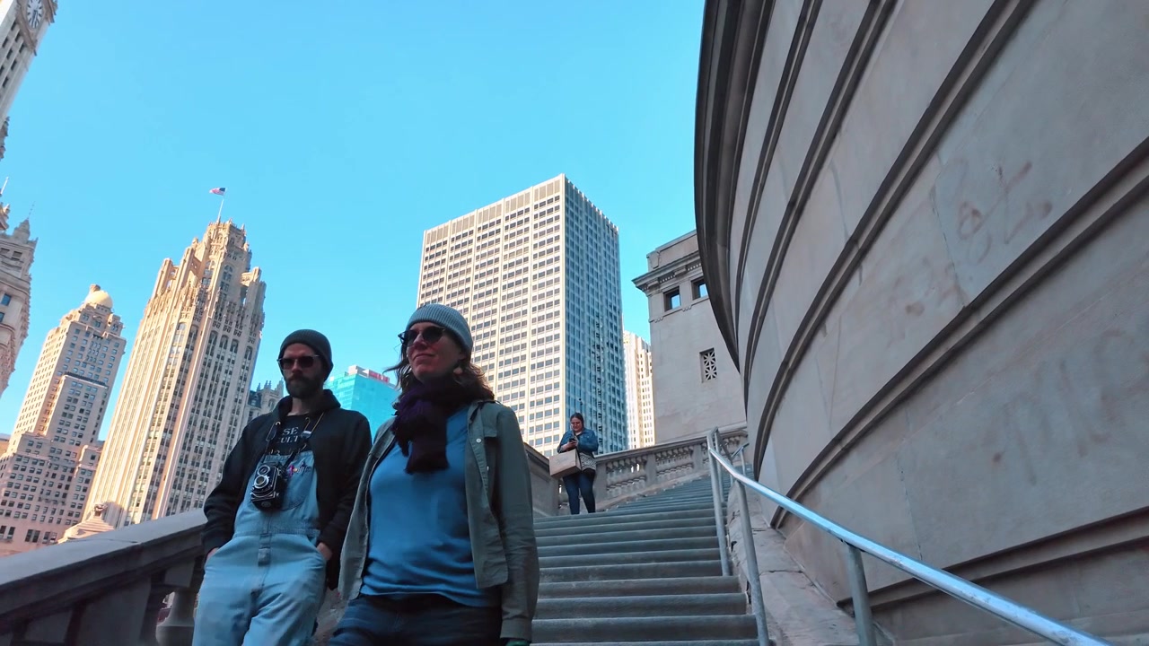 Chicago, USA, 7 maggio 2026: Pedoni che scendono una scalinata in pietra verso la passeggiata lungo il fiume, centro di Chicago. Angolazione bassa di una coppia su gradini intagliati con torri gotiche contro un cielo azzurro e limpido.