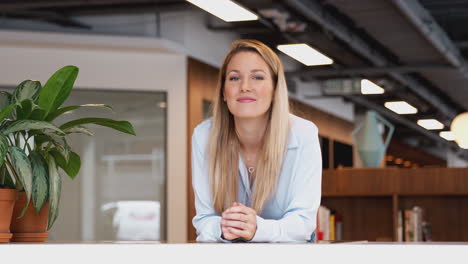 Portrait-Of-Smiling-Casually-Dressed-Young-Businesswoman-Standing-In-Modern-Open-Plan-Workplace