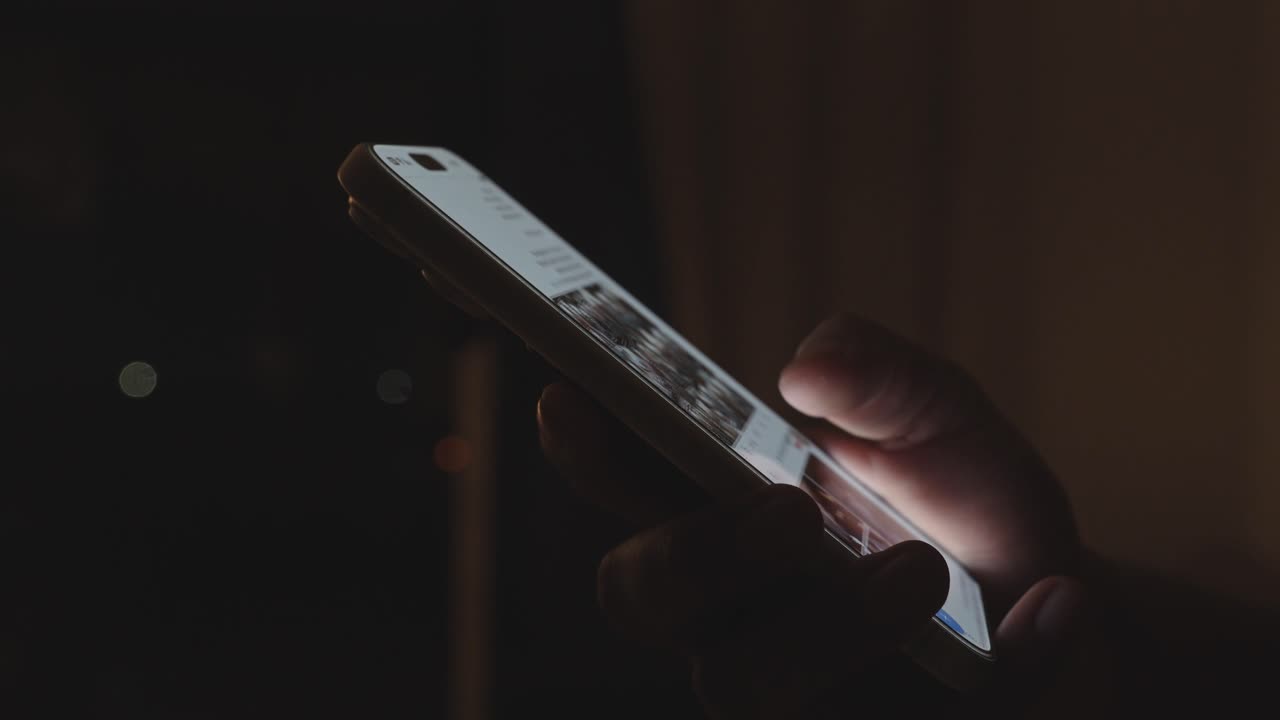 Mano che tiene uno smartphone e scorre un feed di social media in una stanza buia con una scrivania sullo sfondo, illuminata da una luce dai toni caldi. Dipendenza digitale e consumo sconsiderato di contenuti.