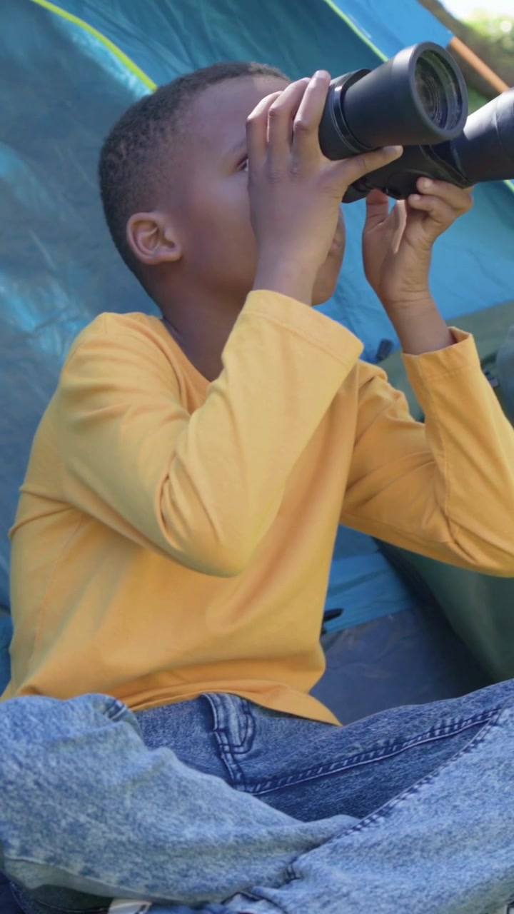 Video verticale: Ragazzo con maglietta color senape che scansiona con binocoli, lasciandoli cadere mentre la telecamera mostra una tenda blu e uno zaino.