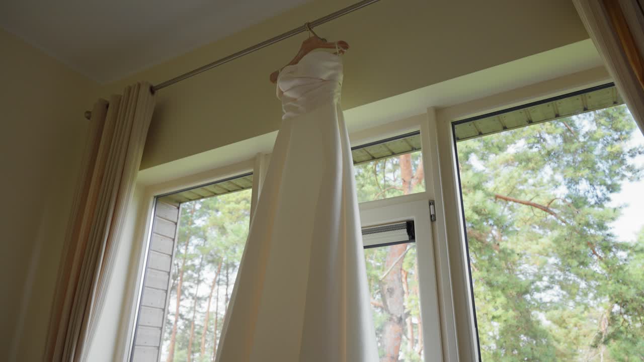 A beautiful, elegant wedding dress hangs gracefully in a sunlit room, casting soft shadows. The morning light enhances its silk texture. Perfect serene, romantic bridal scene