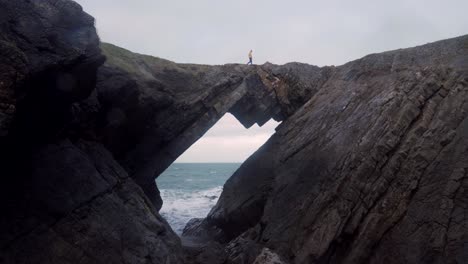 un hombre con una chaqueta amarilla corre a través de una formación rocosa en el puente del diablo, rhossili