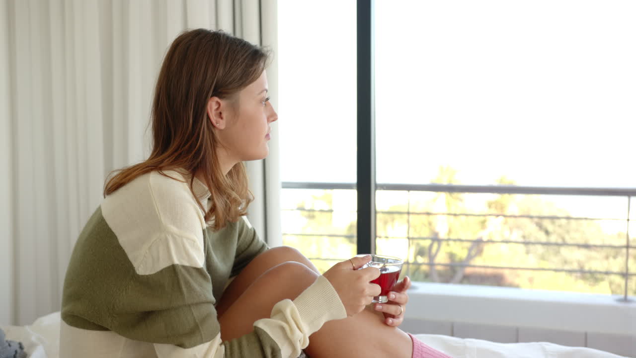 Relaxing in bed, young woman holding cup and looking out window, copy space