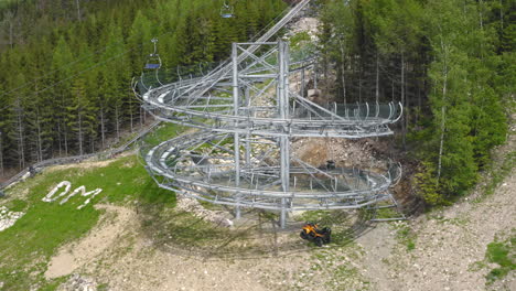 rotating aerial 4k shot of a cart with people spiralling down a big loop of an extreme outdoor roller coaster in the mountains of dolní morava, czech republic with trees around