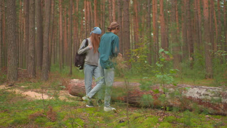 two women approach a fallen tree in lush forest, one, in a green shirt, removes her bag to place it on the tree, while her companion with a blue bandana observes the tree closely