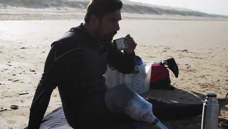 side view a male surfer in wetsuit with artificial leg drinking tea on beach