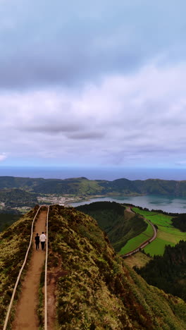 a couple walks by the mountain path leading to the top with observation deck. aerial perspective on the landscape with lake in volcano crater. azores. vertical video.