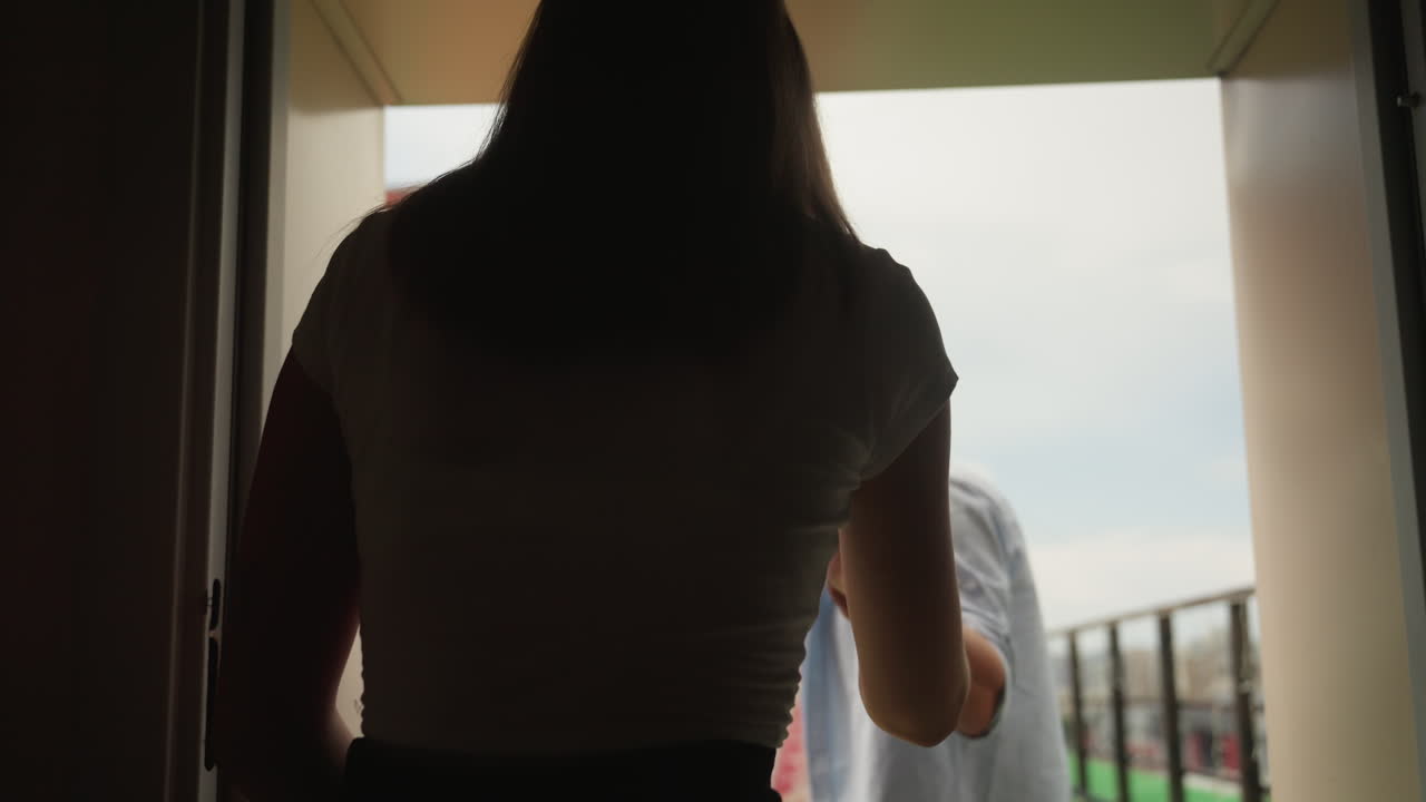 Una coppia esce su un balcone. L'uomo è in testa, con lo skyline della città e un edificio residenziale sullo sfondo. Sul terrazzo c'è una sedia a bolla trasparente. Scena intima diurna: l'uomo si volta indietro sorridendo, la donna lo segue con le mani quasi giunte.
