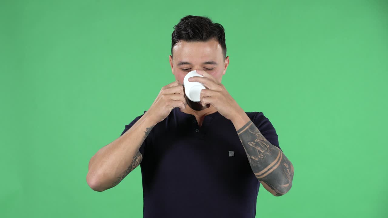 잘생긴 젊은 남자의 초상화는 똑바로 보이고 맛이 나쁜 커피를 마시고 있습니다. 검은 티셔츠를 입고 손에 문신을 한 스타일리시한 머리카락을 가진 갈색을 태우는 스튜디오의 녹색 스크린에서