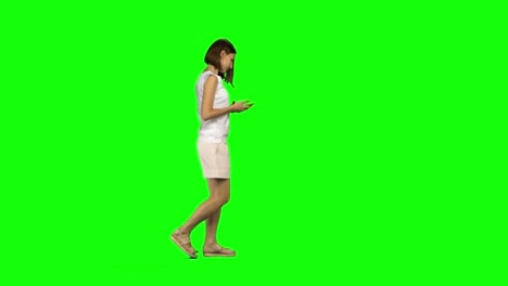 young woman is calmly walking and texting message vie her mobile phone on green screen. profile view