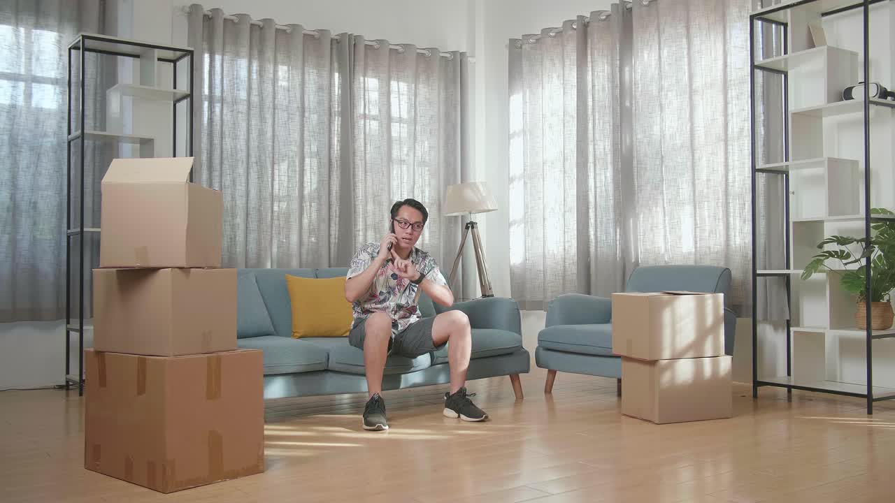 카드보드 상자를 가진 아시아인 남자가 소파에 앉아 새로운 집으로 이사한 후 스마트폰으로 이야기하고 있습니다.