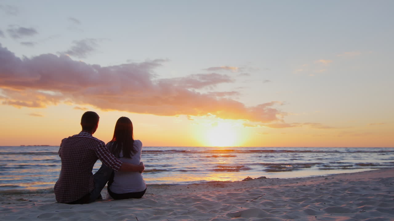 바다에서 일몰을 감상하는 젊은 부부가 모래사장에 껴안고 앉아 prores hq 10비트 비디오