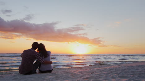 Una-Joven-Pareja-Admirando-La-Atardecer-En-El-Mar,-Sentada-En-Un-Abrazo-En-La-Arena,-Proporciona-Un-Video-Hq-De-10-Bits