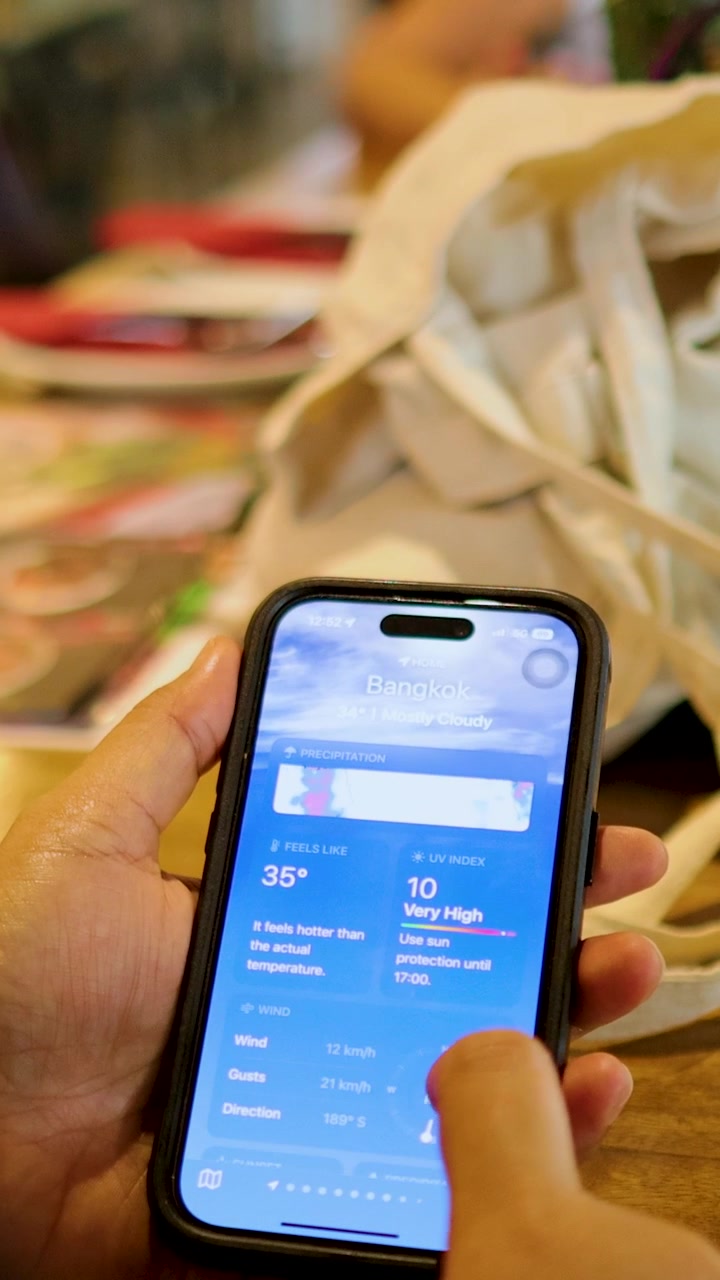 A person interacts with a weather app on a smartphone, checking Bangkok's forecast in a casual indoor setting