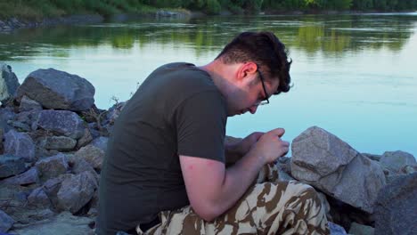 a man in eyeglasses fixing a tangled fishing lure while sitting on rock boulders near the siret river in galati county, romania