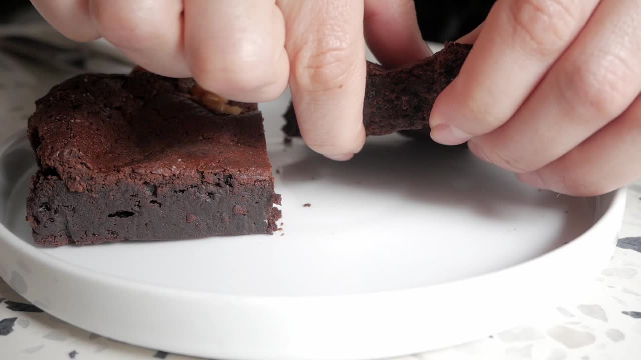 Close up of a hand breaking a chocolate brownie with walnuts on a white plate in a café setting