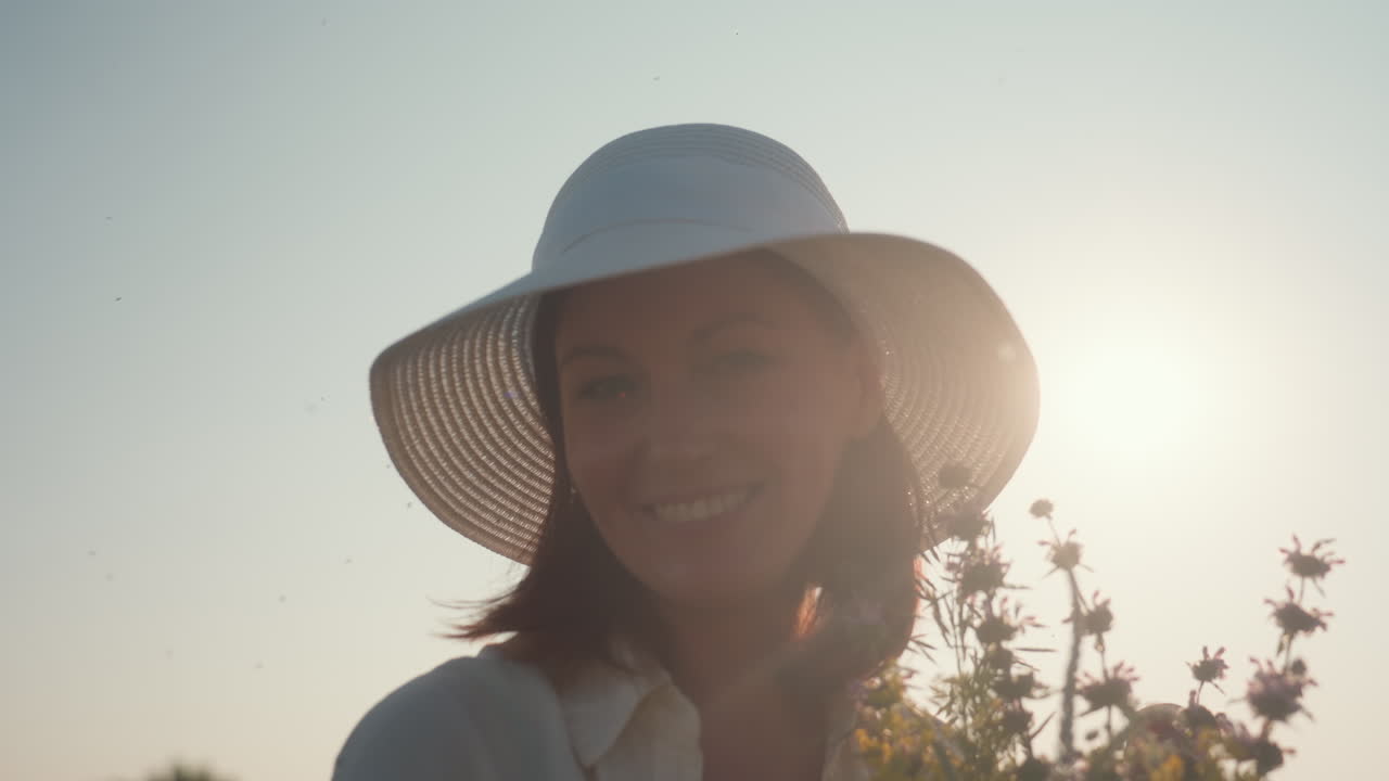 woman in sun hat gently brings wildflowers to nose with closed eyes and warm smile, sunlight illuminating her face, evoking peaceful morning moment surrounded by quiet beauty of countryside