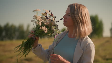 gardener in vast farmland smiling as she takes in scent of freshly picked wildflowers, holding beautiful floral arrangement with distant trees in background, sun gently illuminating environment