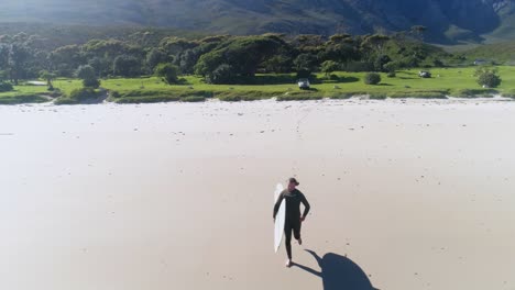 aerial drone shot of a man running with his surfboard on the beach towards the water