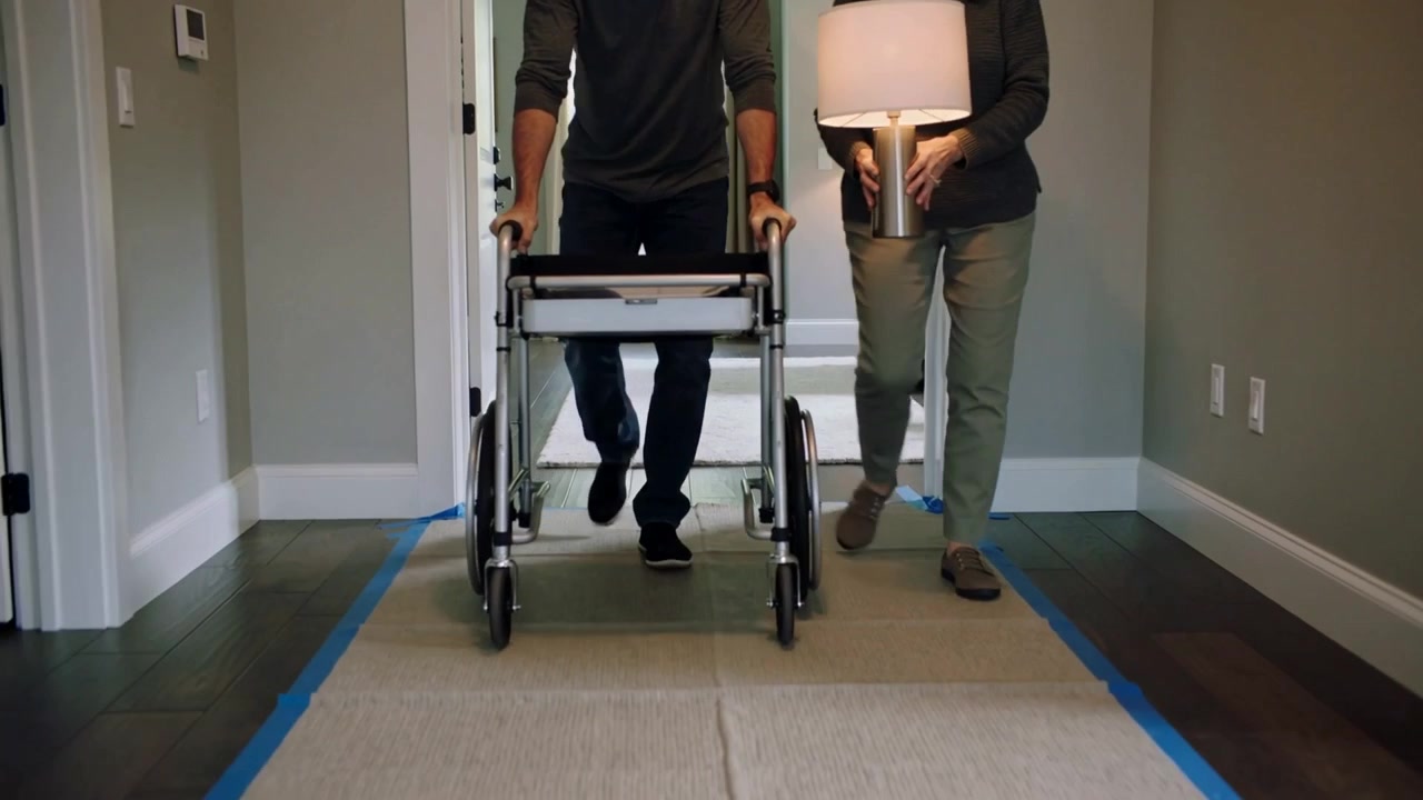 To voksne går fra teppet og styrer en rullator over en teipet løper i gangen, mens de stabiliserer en lampe.