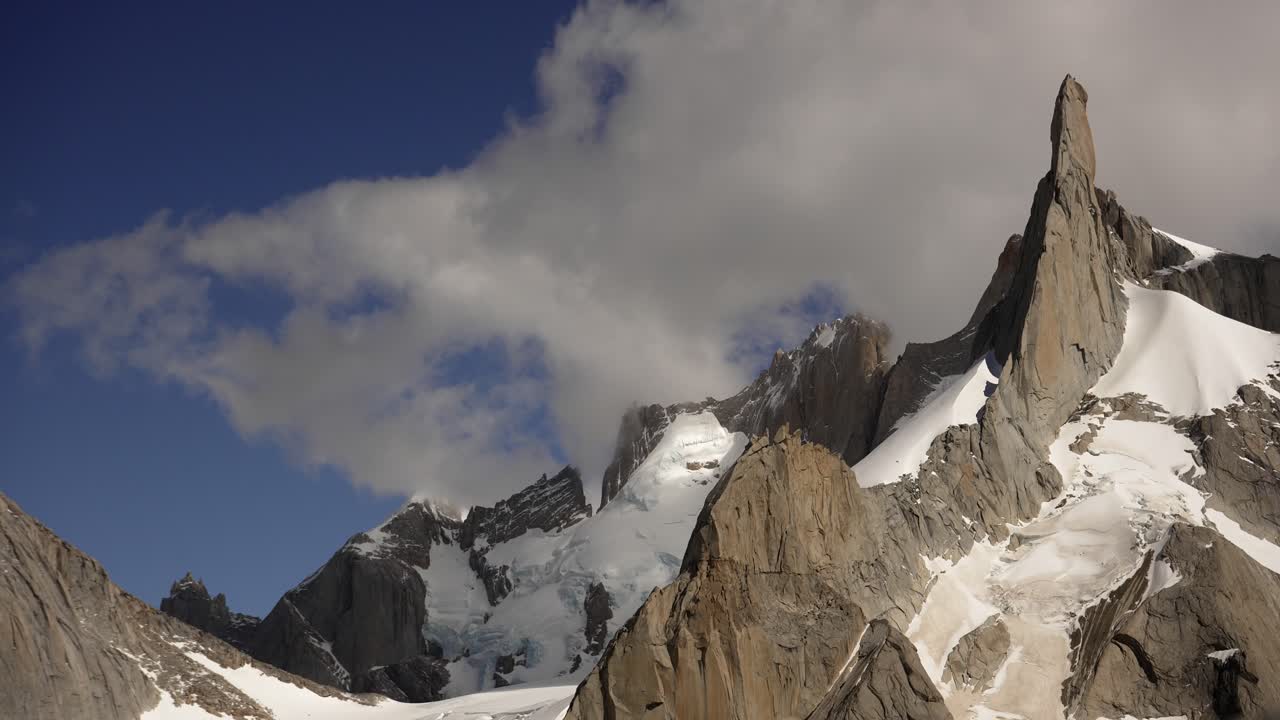 Dynamic timelapse of granite mountains in Patagonia. Clouds race across a deep blue sky above snow-covered peaks, creating a dramatic and rugged alpine scene
