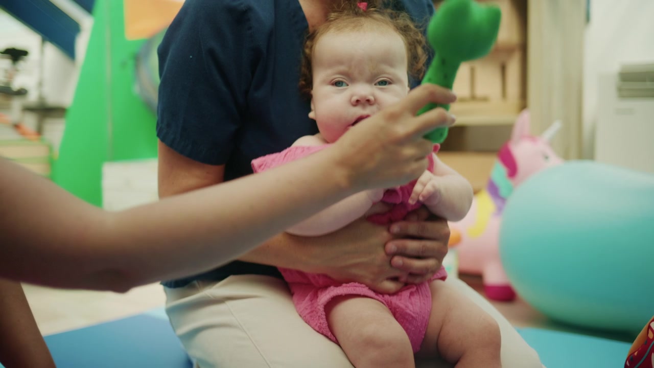 Pediatra ou terapeuta segurando um bebê, oferecendo suporte durante uma sessão de reabilitação com brinquedos e equipamentos de exercício