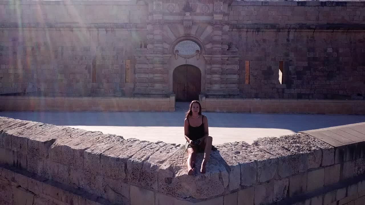 Aerial drone shot of a beautiful, caucasian woman flying and filming herself while she is sitting on the walls of historical Fort Manoel on Manoel Island, Malta. Drone flying backwards.