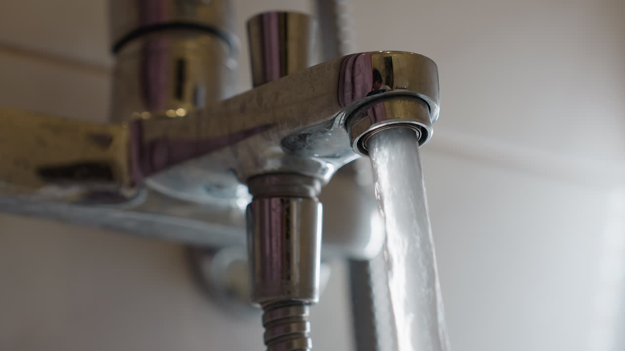 Close up of water streaming from chrome bathroom tap with reflections on shiny surface, showing detail of plumbing, sanitation, hygiene, water flow and household usage